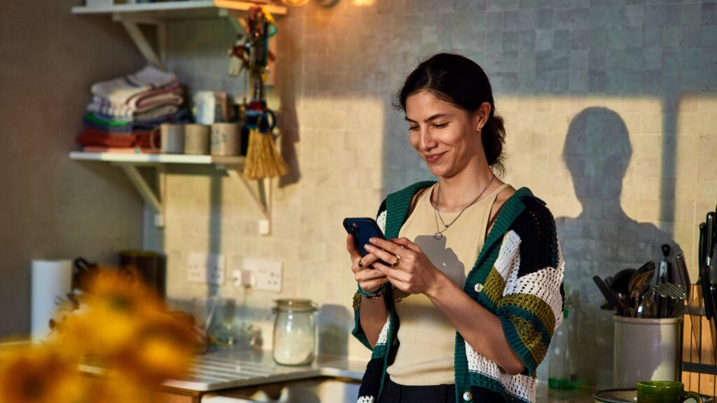 Lächelnde Frau steht in der Küche und schaut auf ihr Smartphone.
